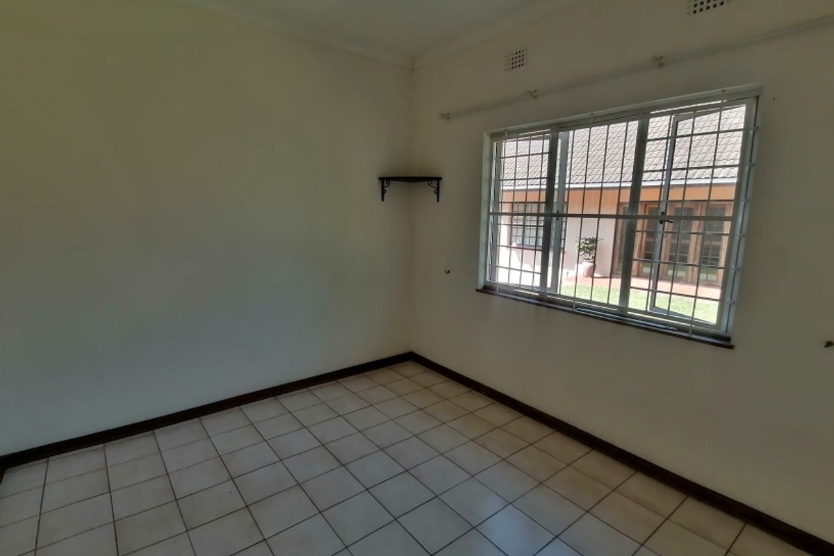 A vacant bedroom featuring tiled flooring, a ceiling fan, and a large window with security bars overlooking the rear garden area.