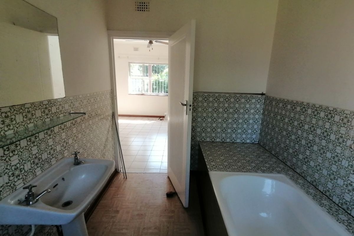 A dated bathroom featuring a pedestal sink, built-in bathtub, and patterned wall tiles, with an adjacent view into a tiled living area.