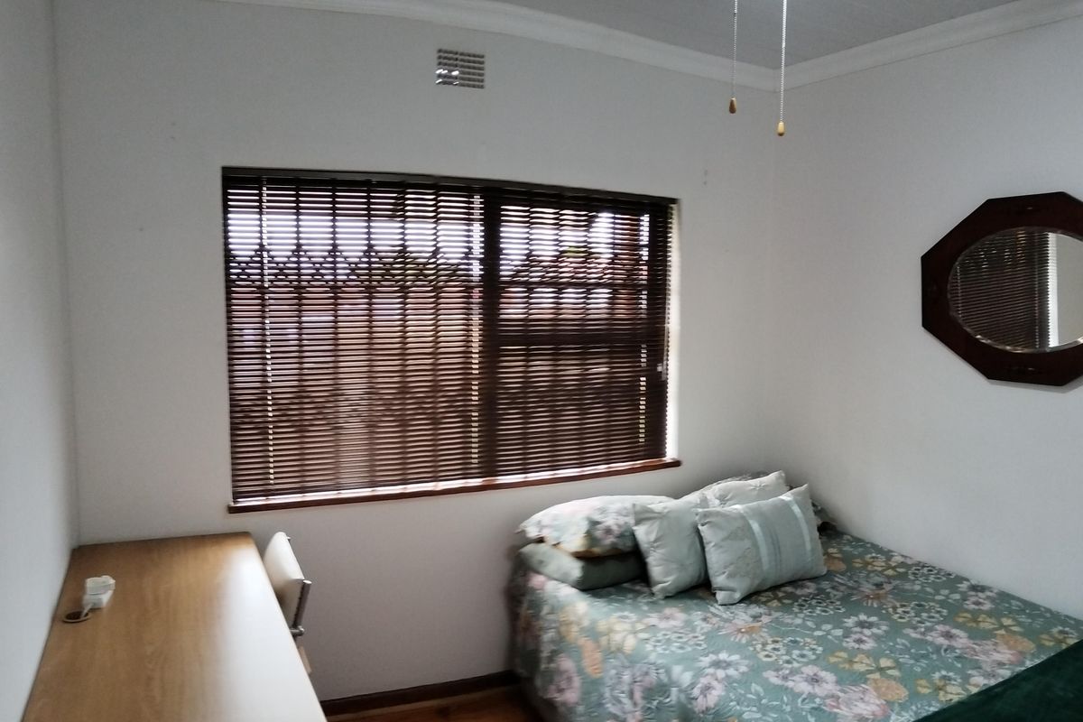 A bedroom featuring timber flooring, a ceiling fan with integrated lighting, and a window fitted with security bars and blinds.