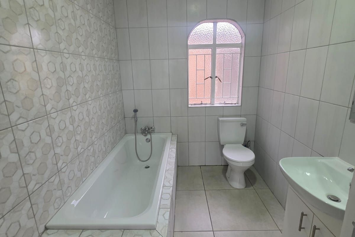 A functional bathroom featuring a built-in bathtub with shower mixer, a pedestal vanity, and a toilet, finished with full-height wall tiling.