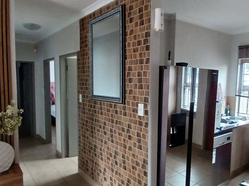 An open-plan living area featuring a decorative tiled feature wall, tiled flooring, and a compact kitchen with built-in cabinetry and window blinds.