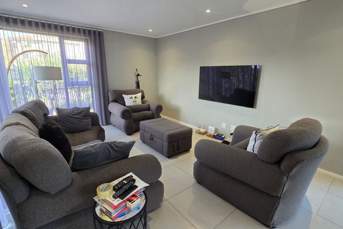 A comfortable living room featuring neutral tiled flooring, recessed ceiling lighting, and a wall-mounted television, with large windows providing natural light.