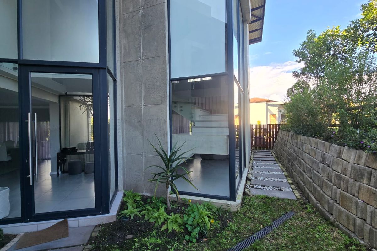 Modern exterior view featuring floor-to-ceiling glass windows, concrete panel cladding, and a paved side walkway with a stone retaining wall.