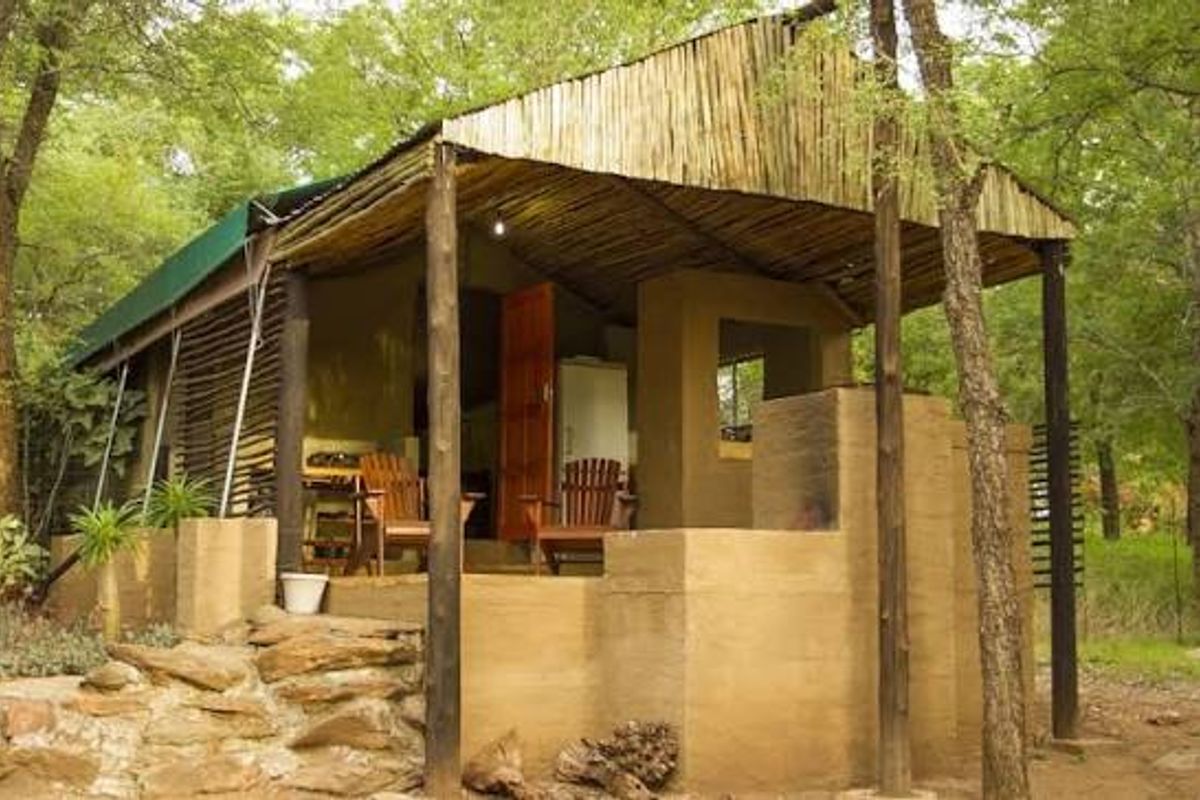 A rustic farm-style cabin featuring a covered patio, natural stone foundation, and traditional thatched roof structure nestled in a wooded setting.