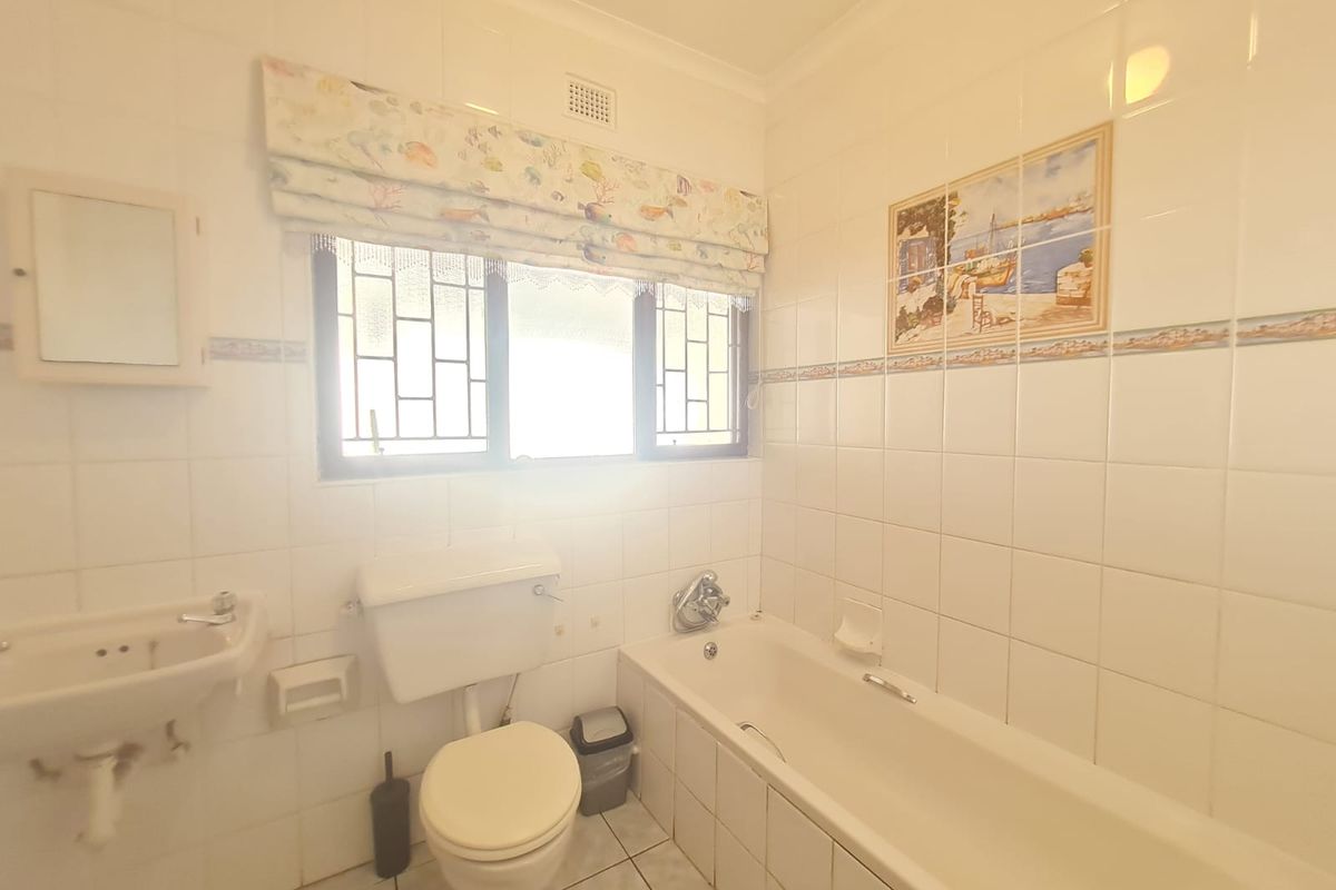 A functional bathroom featuring a standard bathtub, wall-mounted basin, and toilet, finished with classic white wall tiles and a window with security bars.