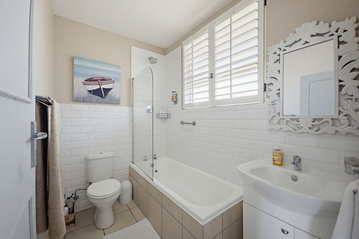 A well-maintained bathroom featuring a white bathtub with a glass shower screen, subway tile backsplash, vanity basin, and white plantation shutters.