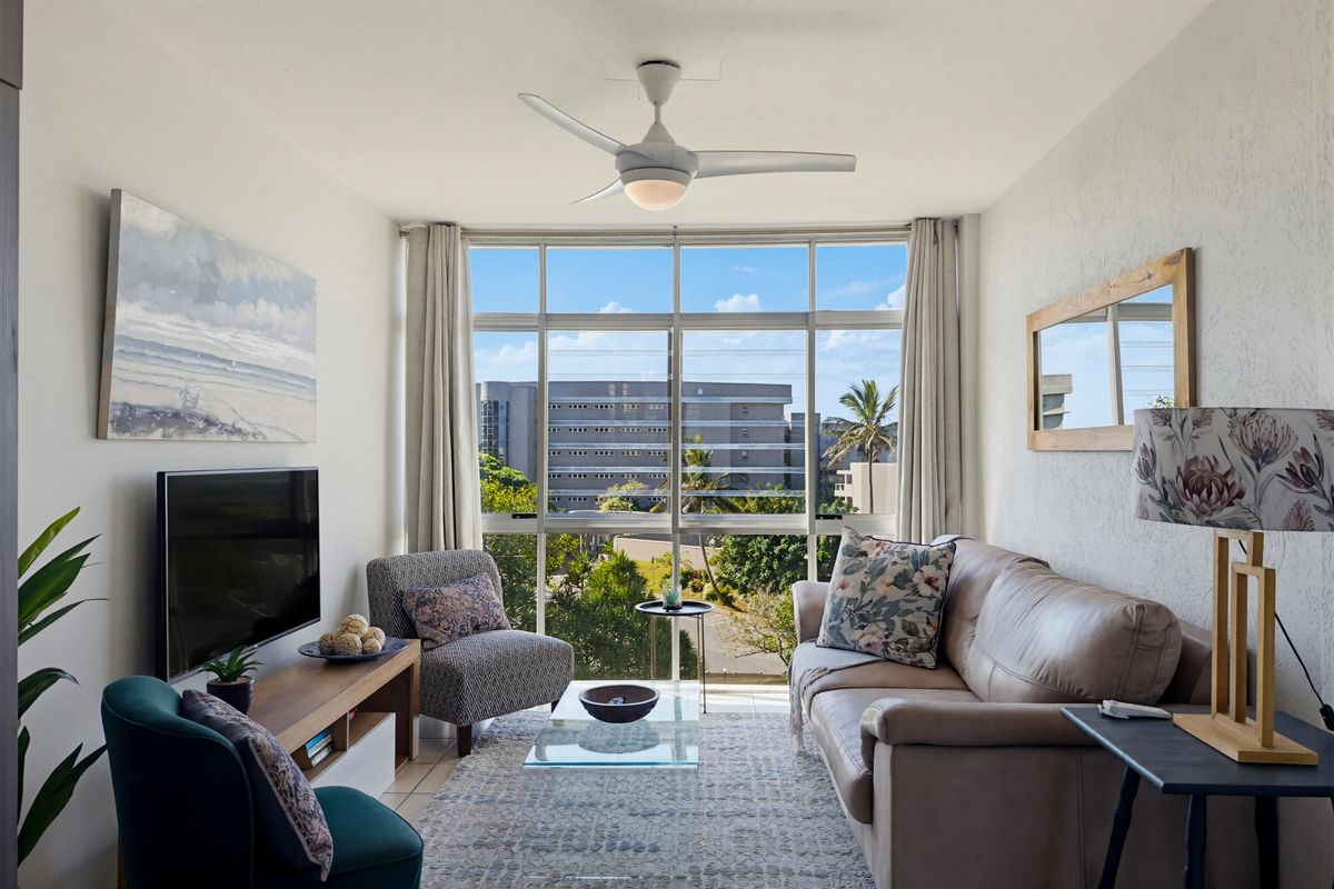 A bright living room featuring large floor-to-ceiling windows with an urban view, a ceiling fan, and neutral wall finishes.