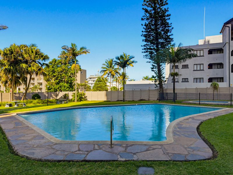A communal swimming pool area featuring a paved surround, manicured lawn, and established palm trees within a secure complex setting.