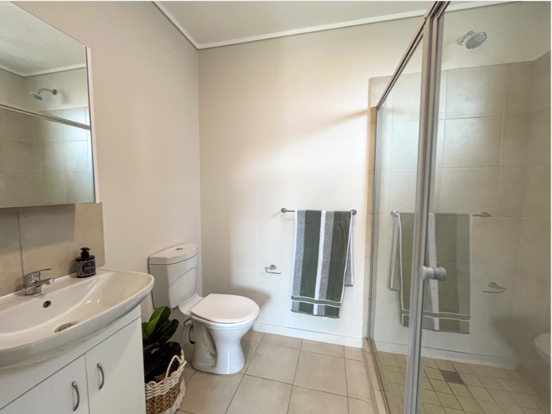 A clean and functional bathroom featuring a vanity cabinet, toilet, and a glass-enclosed shower with neutral wall and floor tiling.