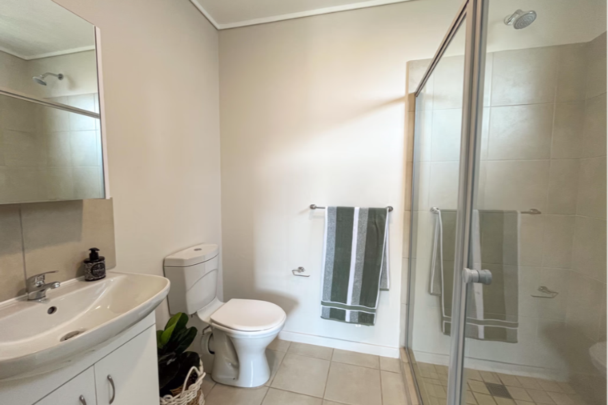 A clean and functional bathroom featuring a vanity cabinet, toilet, and a glass-enclosed shower with neutral wall and floor tiling.