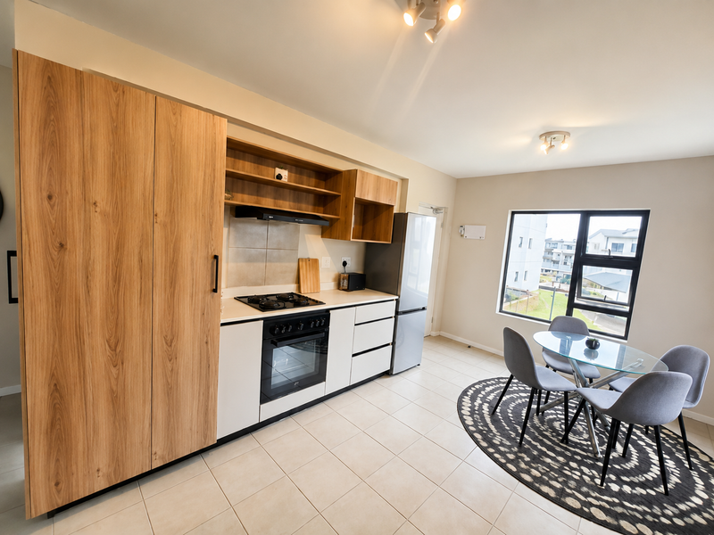 Modern open-plan kitchen and dining area featuring light wood-finish cabinetry, built-in appliances, tiled flooring, and large windows providing natural light.