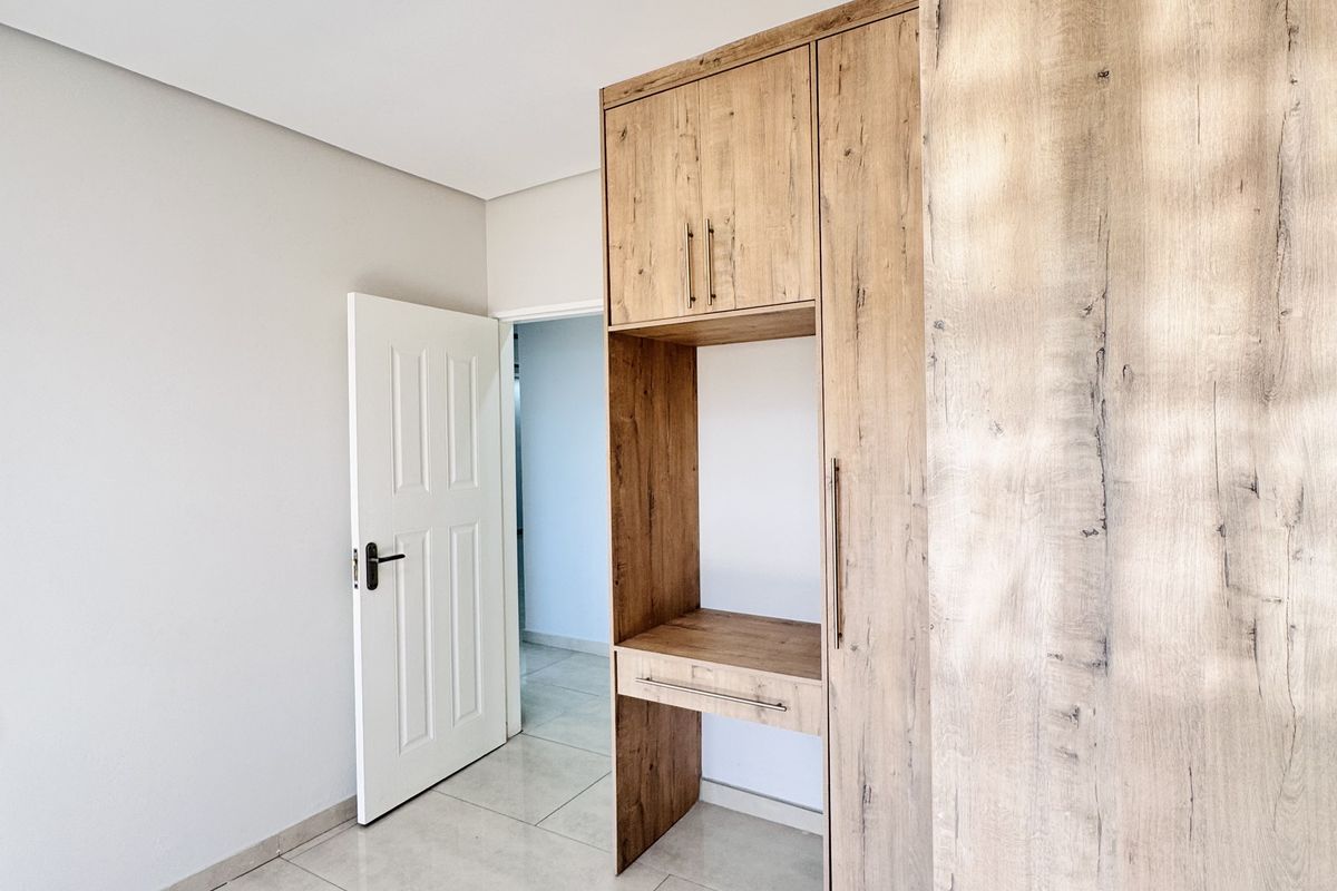 A bright bedroom featuring modern wood-grain built-in cabinetry with a desk area and neutral tiled flooring.