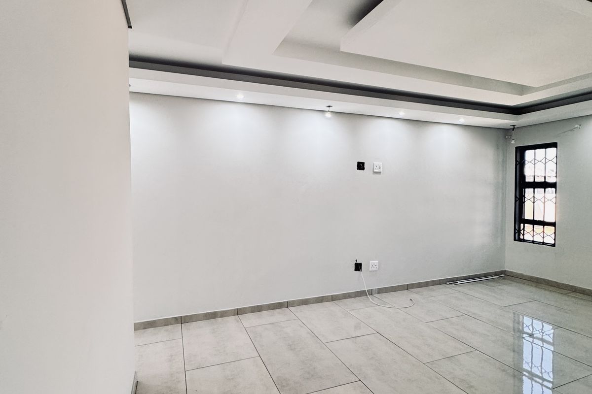 A bright, empty living room featuring modern large-format floor tiles, a decorative recessed tray ceiling with integrated lighting, and a security-grilled window.