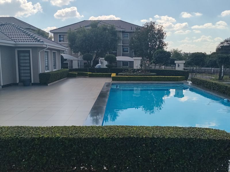 A well-maintained outdoor pool area featuring a large swimming pool, spacious tiled patio, and manicured garden hedging.