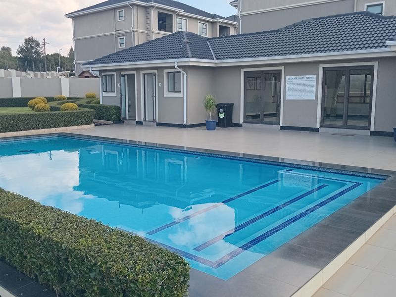 A well-maintained communal swimming pool area featuring a tiled deck, landscaped hedges, and access to the adjacent building structure.