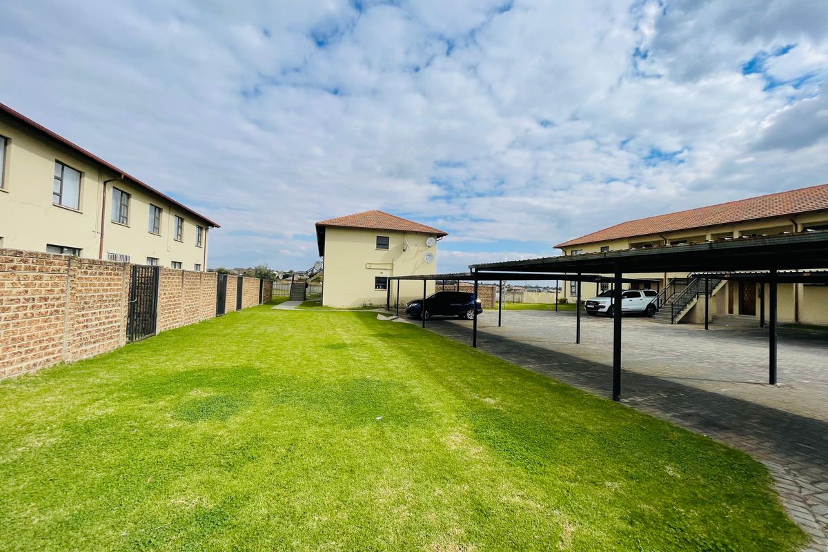 Exterior view of the apartment complex grounds featuring a paved parking area with covered carports, a brick boundary wall, and a well-maintained lawn.