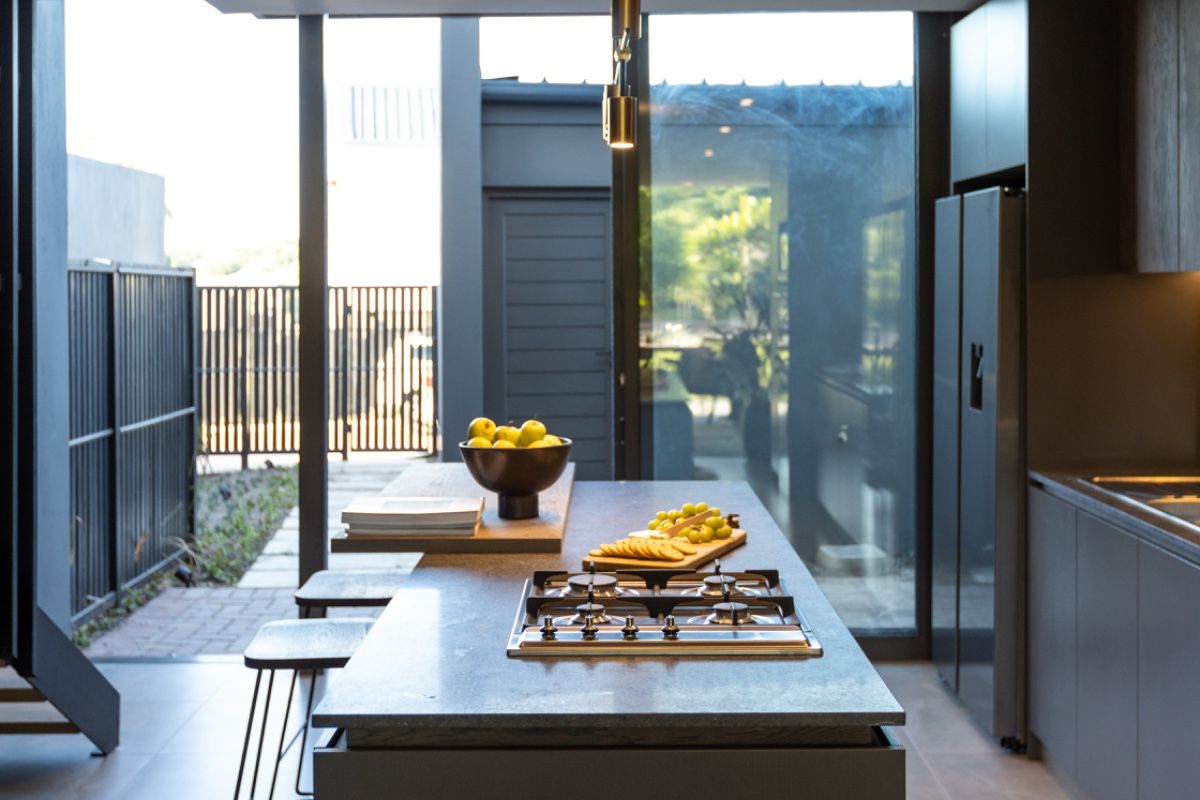 Modern open-plan kitchen featuring a central island with a built-in gas hob, sleek cabinetry, and floor-to-ceiling glass doors opening to an outdoor area.