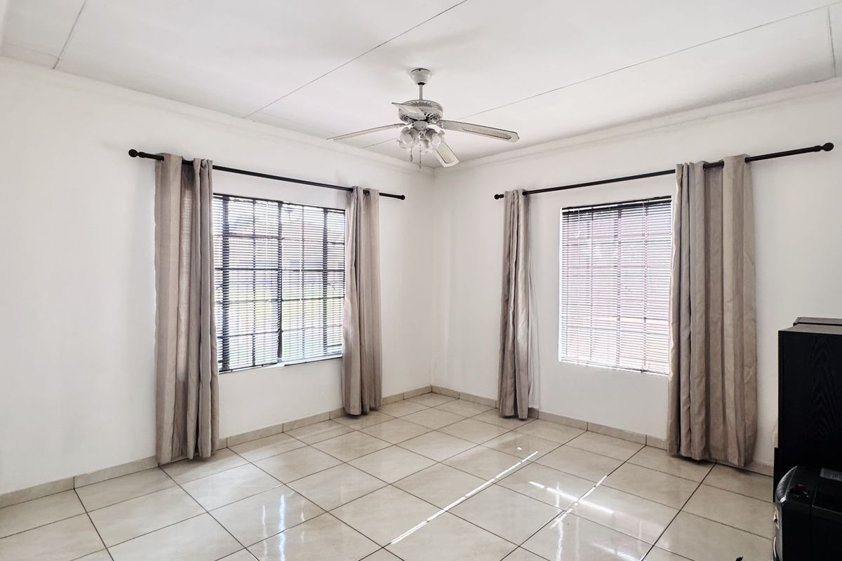 A bright and spacious living room featuring neutral floor tiles, a ceiling fan, and large windows with fitted blinds.