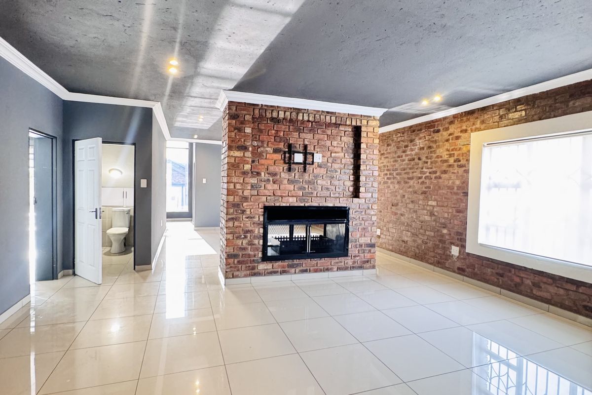 Spacious living area featuring a central exposed brick fireplace, polished porcelain floor tiles, and a view into an adjacent bathroom.