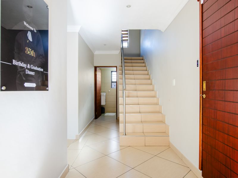 A bright and clean entrance hallway featuring tiled flooring, a staircase with a stainless steel handrail, and access to an adjacent room.