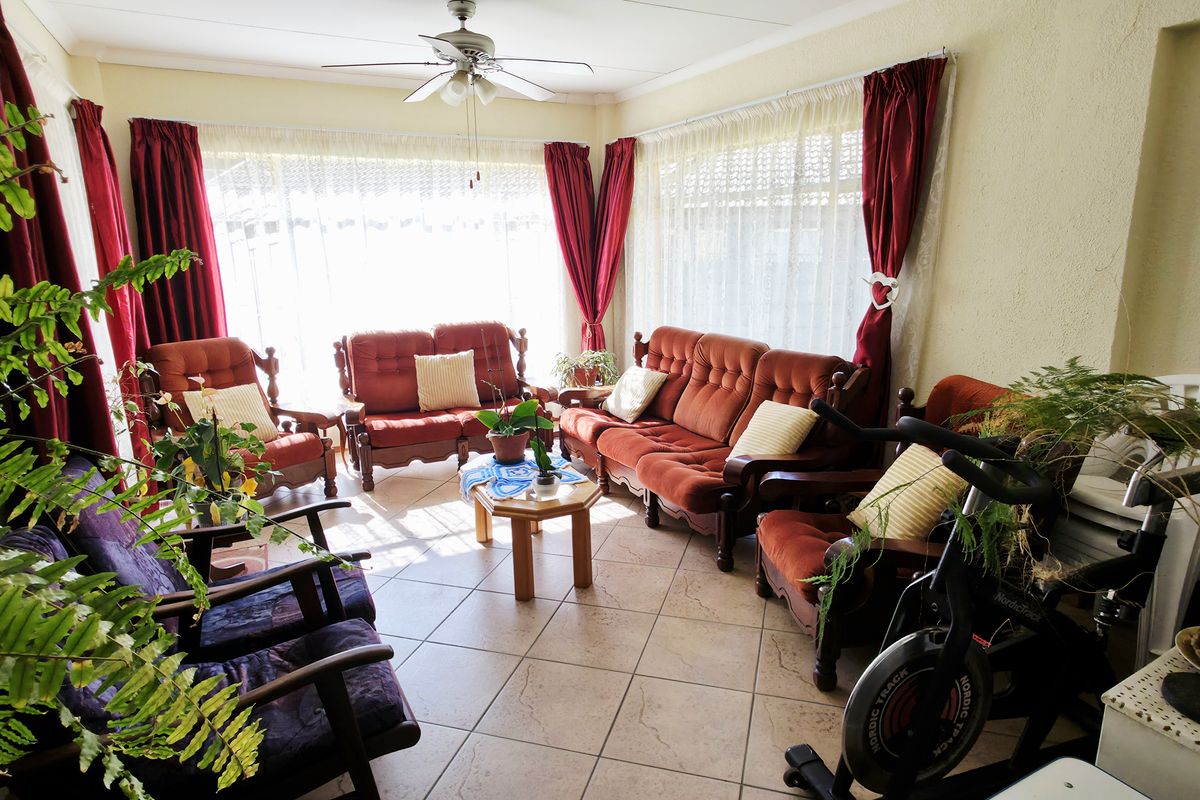 A sunlit living room featuring tiled flooring, a ceiling fan, and large windows with curtains, providing a bright and comfortable space.