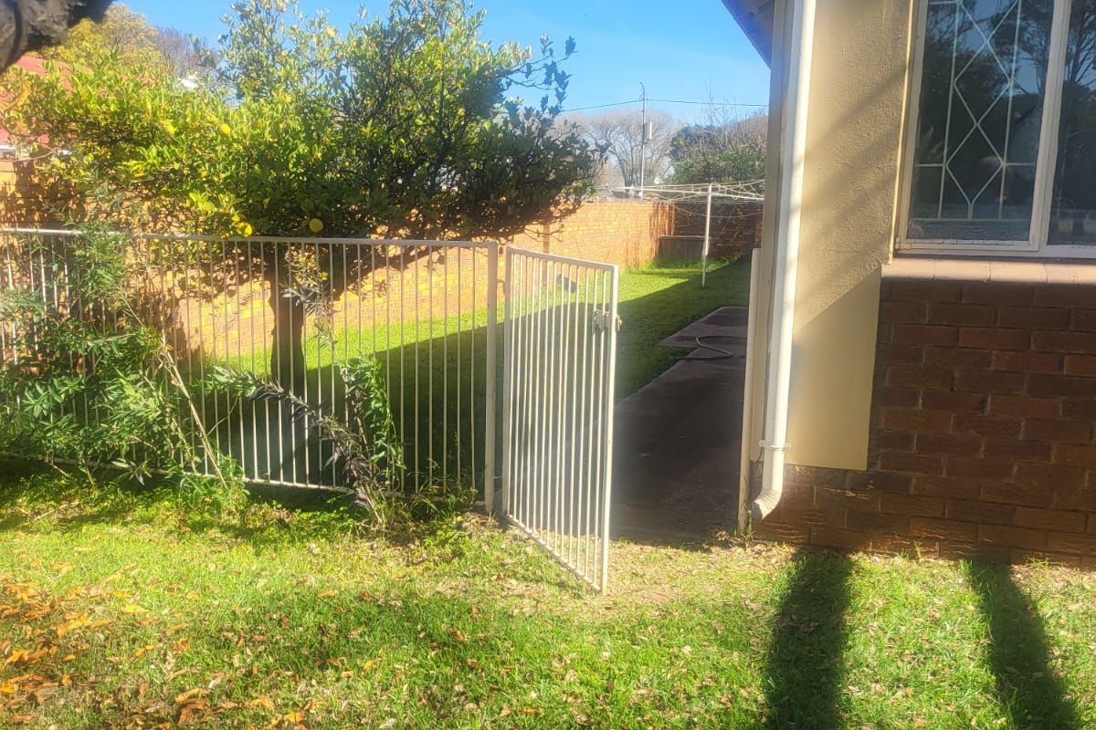 A view of the rear garden area featuring a lawn, a metal security gate, and the side elevation of the house with brick and plaster finishes.