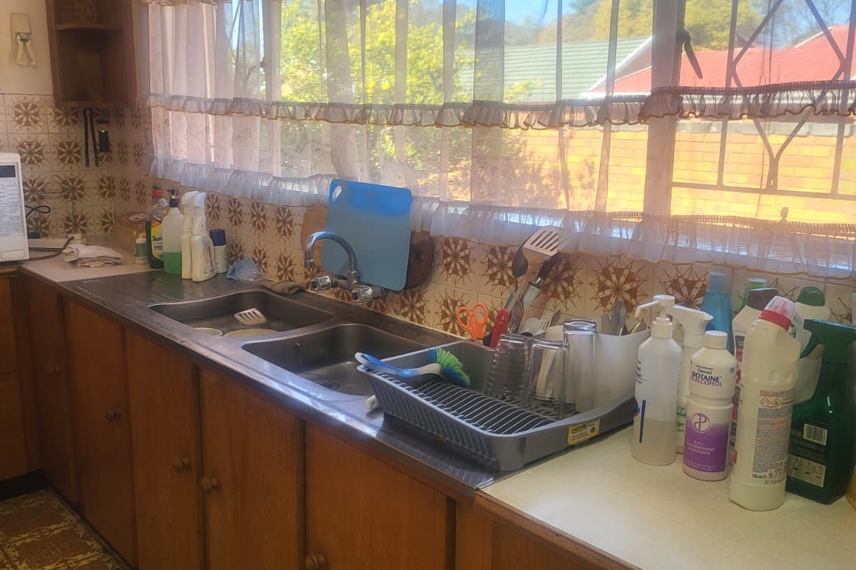 A dated kitchen featuring original wooden cabinetry, patterned wall tiles, and a double stainless steel sink under a large window.