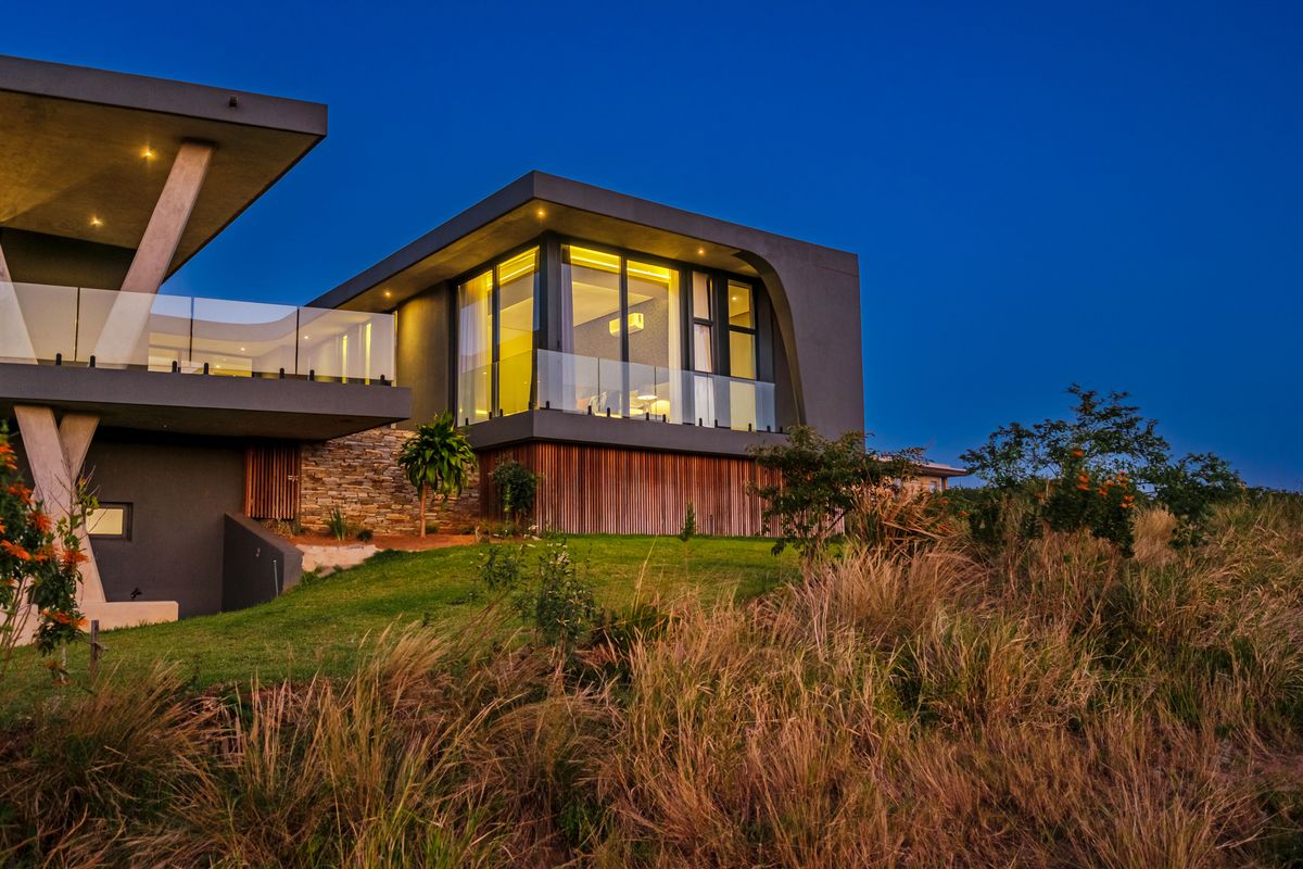 Modern architectural rear elevation featuring stone and timber cladding, glass balustrades, and expansive windows set against a landscaped garden at twilight.