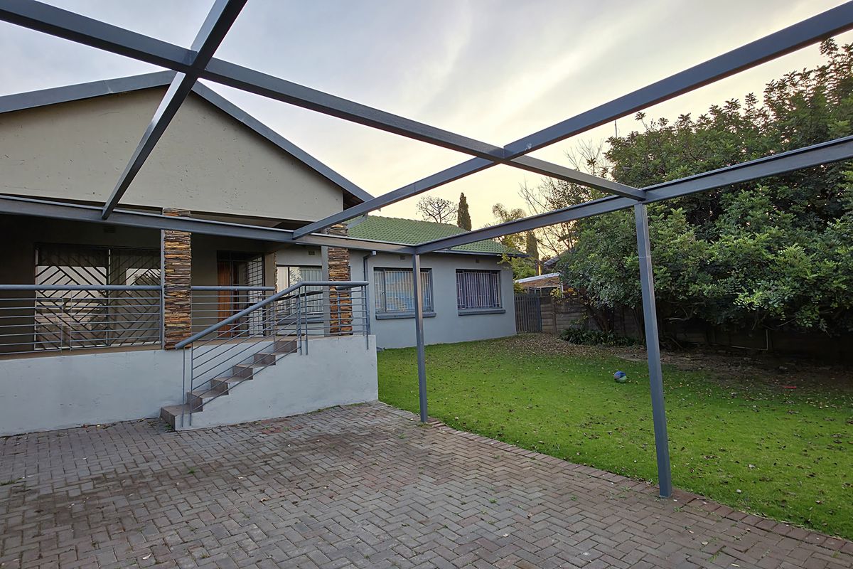 Rear exterior view featuring a paved patio area with a metal pergola structure, stone-clad pillars, and a well-maintained garden.