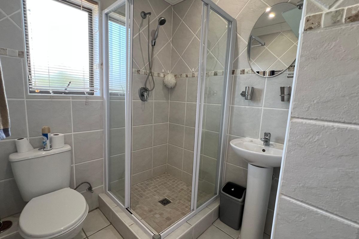 A functional bathroom featuring a corner shower enclosure, pedestal basin, and toilet with neutral wall tiling and natural light from a window.
