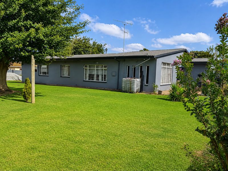 A view of the rear exterior of the house featuring a well-maintained lawn, mature trees, and a grey painted facade with aluminium windows.