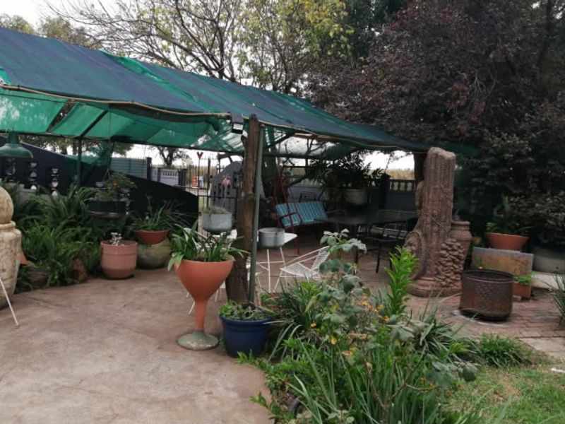 A covered outdoor patio area featuring paved flooring and a shade-net structure supported by timber posts, set within a garden environment.