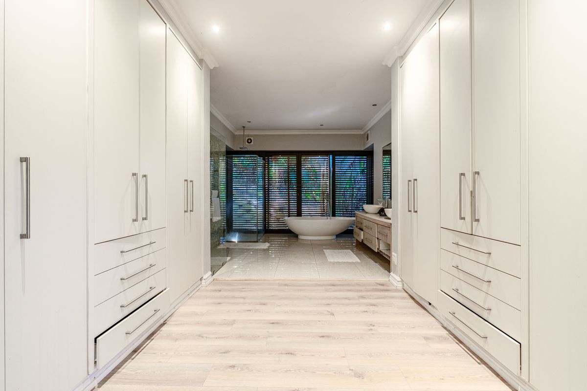 Spacious modern bathroom featuring extensive built-in cabinetry, a central freestanding bathtub, double vanity, and a walk-in shower with natural light through shutter blinds.