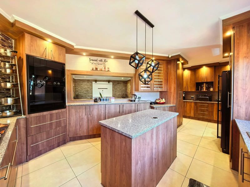 A well-appointed kitchen featuring warm wood-toned cabinetry, granite countertops, a central island, and modern pendant lighting with a convenient pass-through window.