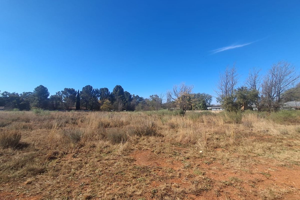 A wide-angle view of a vacant residential plot featuring natural grass and scattered trees under a clear blue sky.