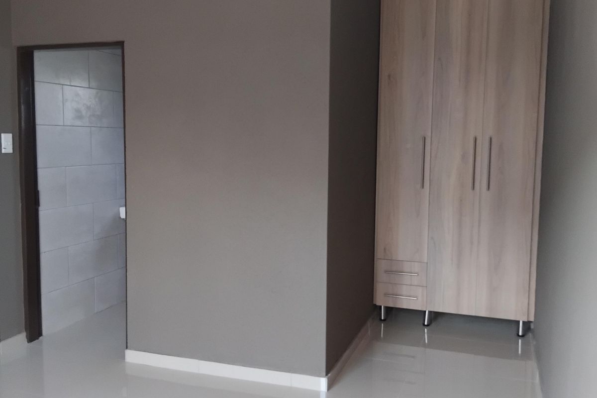 A clean, empty bedroom featuring neutral grey walls, light wood-finish built-in wardrobes, recessed ceiling lighting, and polished floor tiles.