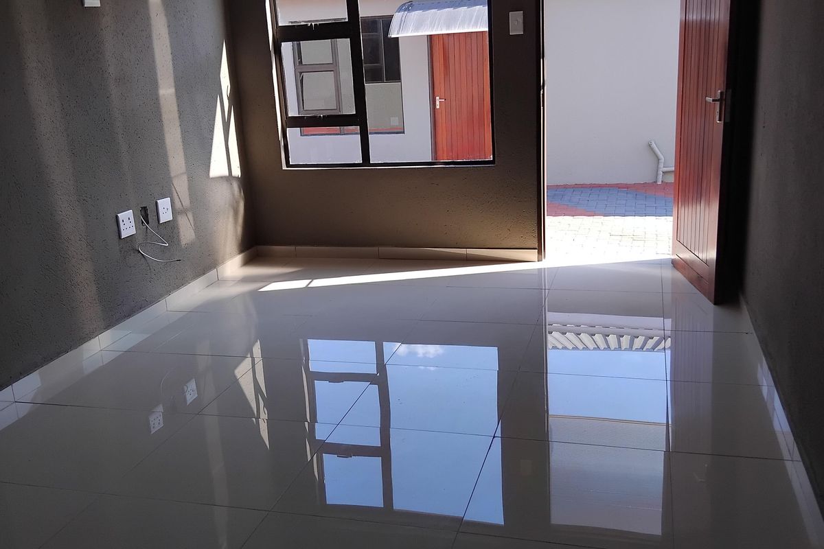 A bright, empty living room featuring polished floor tiles, recessed downlighting, and direct access to an outdoor area through a timber door.