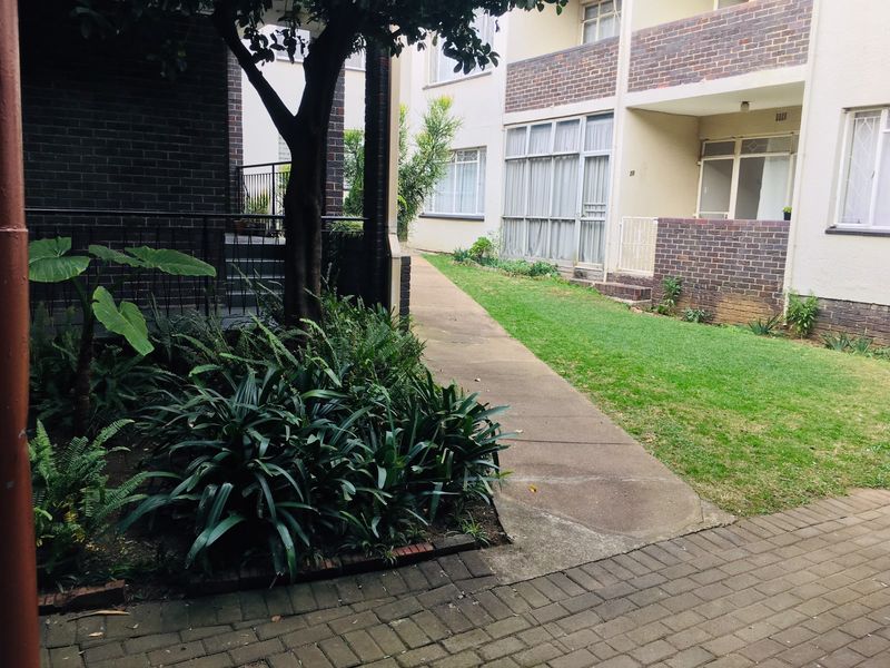 A view of the communal garden area featuring a concrete pathway, established landscaping, and brick facade of the apartment building.