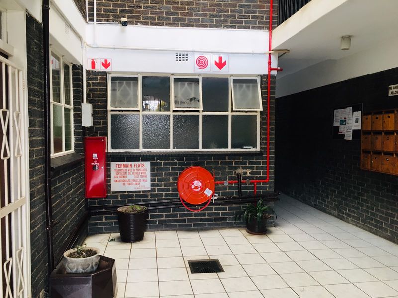 A communal exterior courtyard area featuring brick walls, tiled flooring, fire safety equipment, and wall-mounted mailboxes for the flat complex.