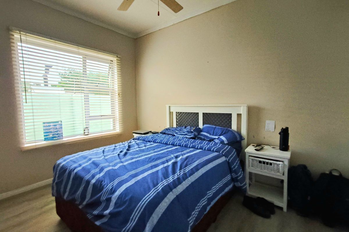 A functional bedroom featuring a ceiling fan, light-toned flooring, and a large window with fitted blinds, providing natural light.