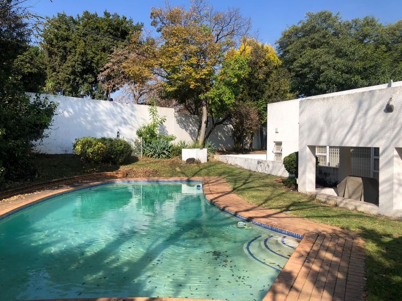 A private backyard featuring a swimming pool with brick coping, surrounded by a lawn and mature trees, adjacent to a covered patio area.