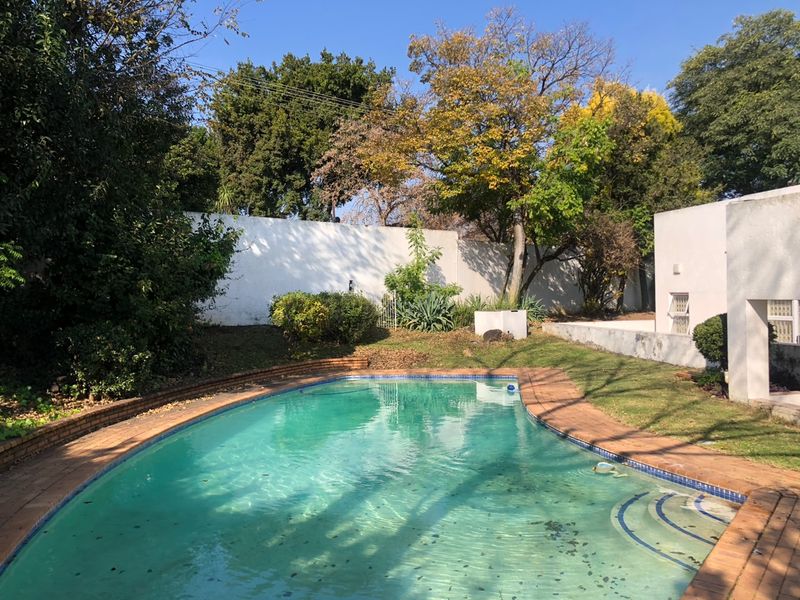 A private outdoor pool area featuring a curved swimming pool with brick coping, surrounded by established garden landscaping and a perimeter boundary wall.
