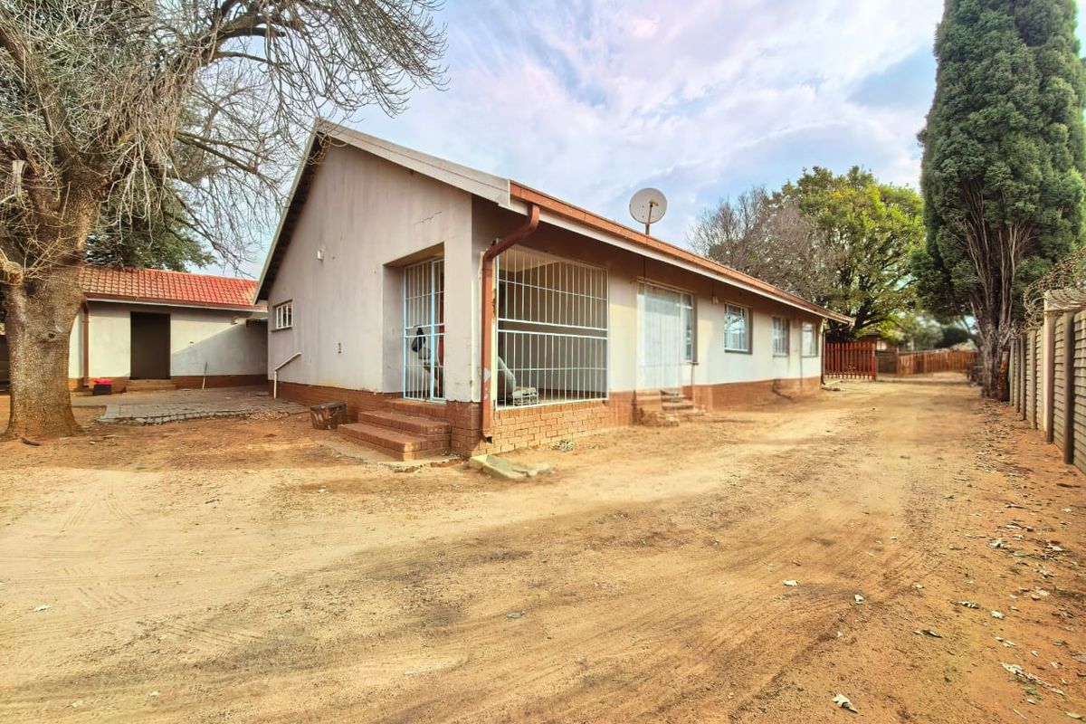 Exterior view of a single-story house featuring a tiled roof, security bars on windows, and a large unpaved yard area.