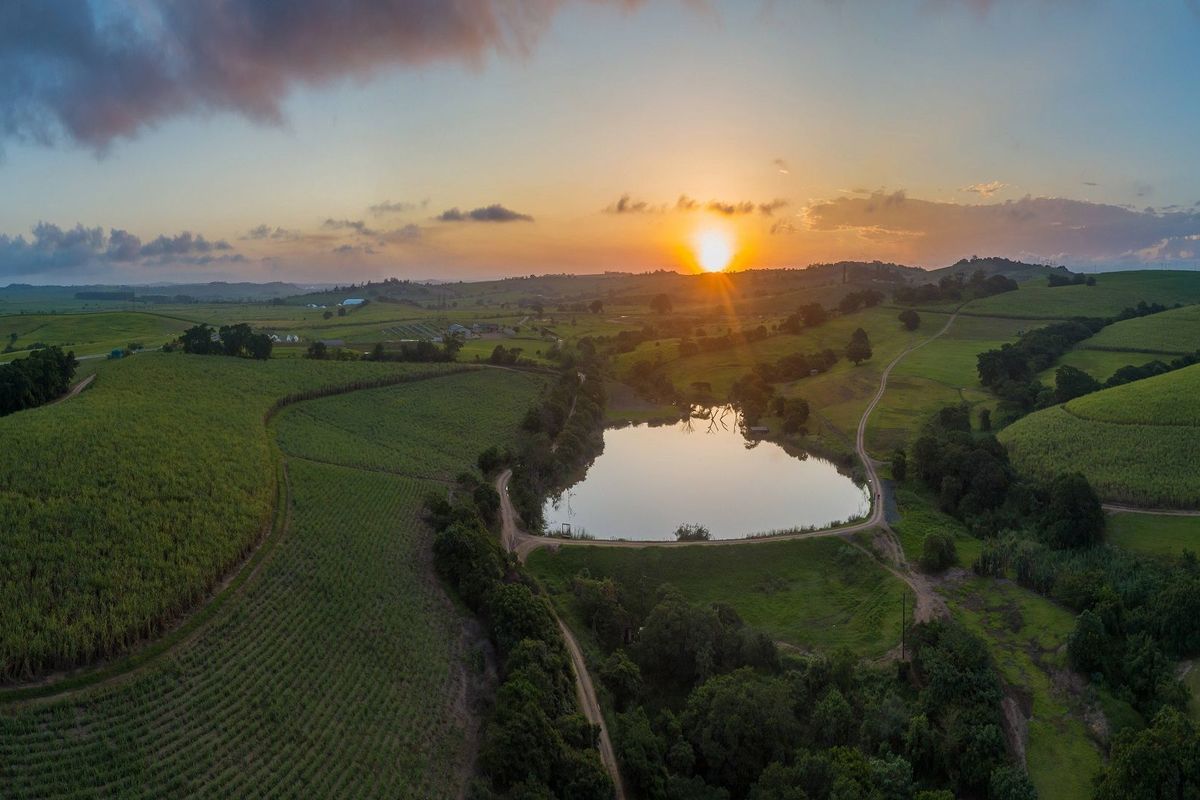 An expansive aerial view of the vacant residential land featuring rolling green hills, agricultural fields, and a central dam at sunset.