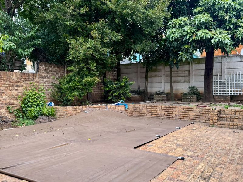 Outdoor pool area featuring a brick-paved patio, retaining walls, and a covered swimming pool surrounded by mature trees and boundary walls.