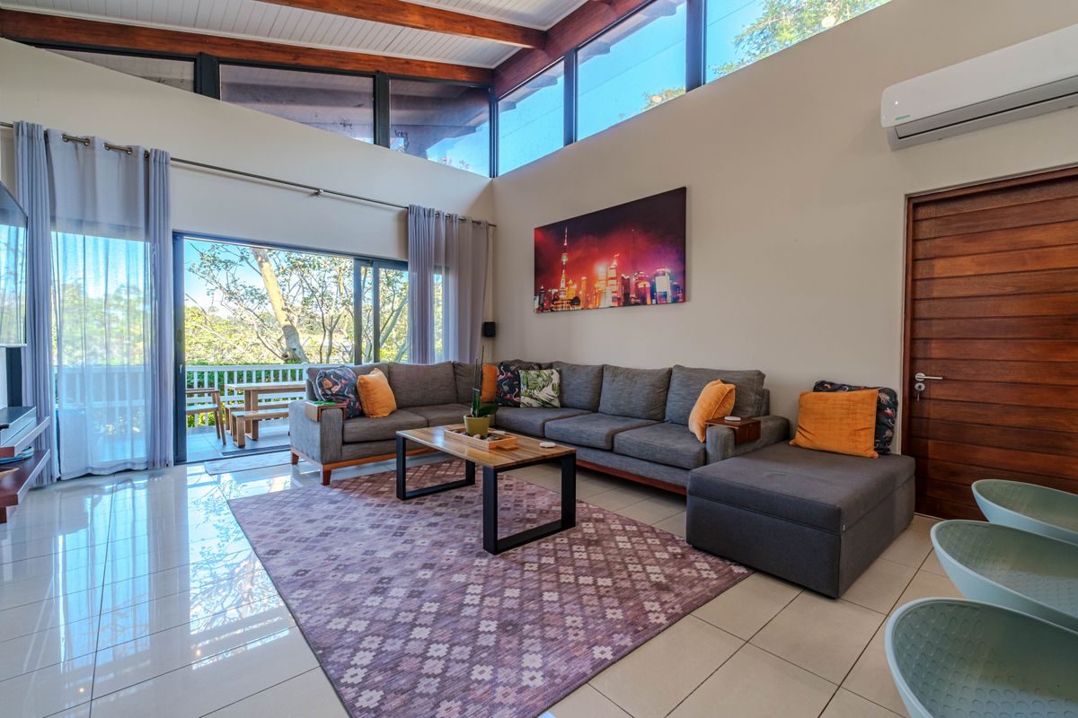 Spacious living room with high vaulted ceilings, exposed timber beams, large clerestory windows, and sliding glass doors opening to an outdoor patio area.