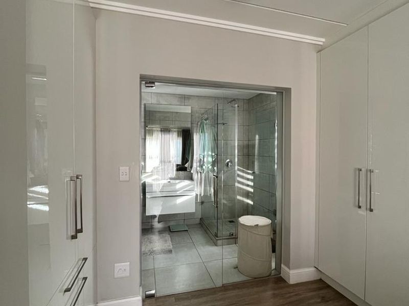 A modern dressing area featuring extensive built-in high-gloss cabinetry, leading through a glass door into a contemporary en-suite bathroom with tiled finishes.
