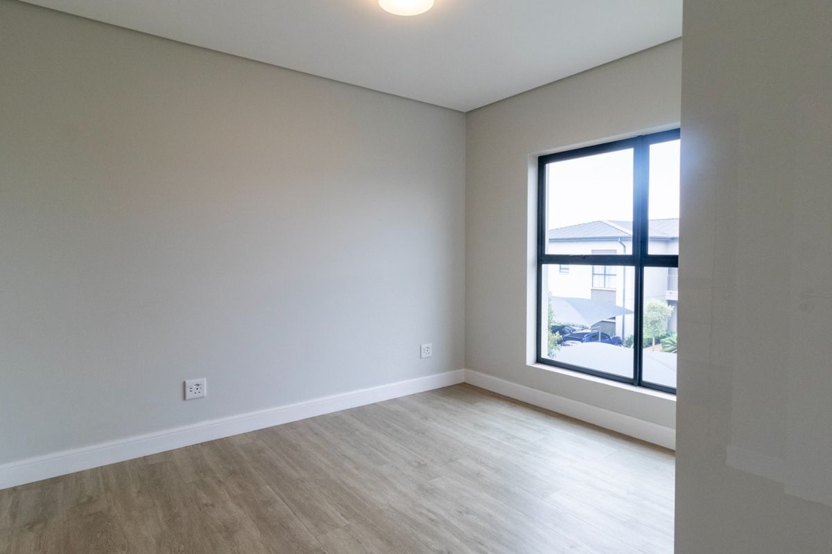 A bright, empty bedroom featuring modern light wood-look flooring, neutral wall paint, and a large window with black framing.