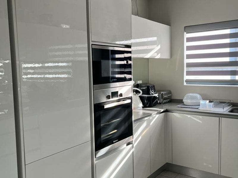 Modern kitchen featuring high-gloss white cabinetry, integrated oven and microwave, tiled flooring, and a window with fitted blinds.