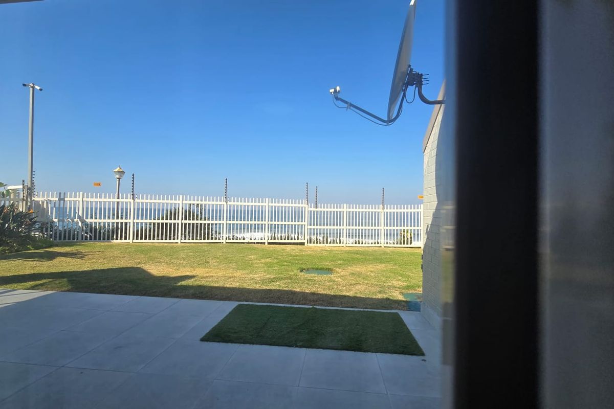 A view of the private rear garden featuring a paved patio area, lawn, and secure perimeter fencing with ocean views in the distance.